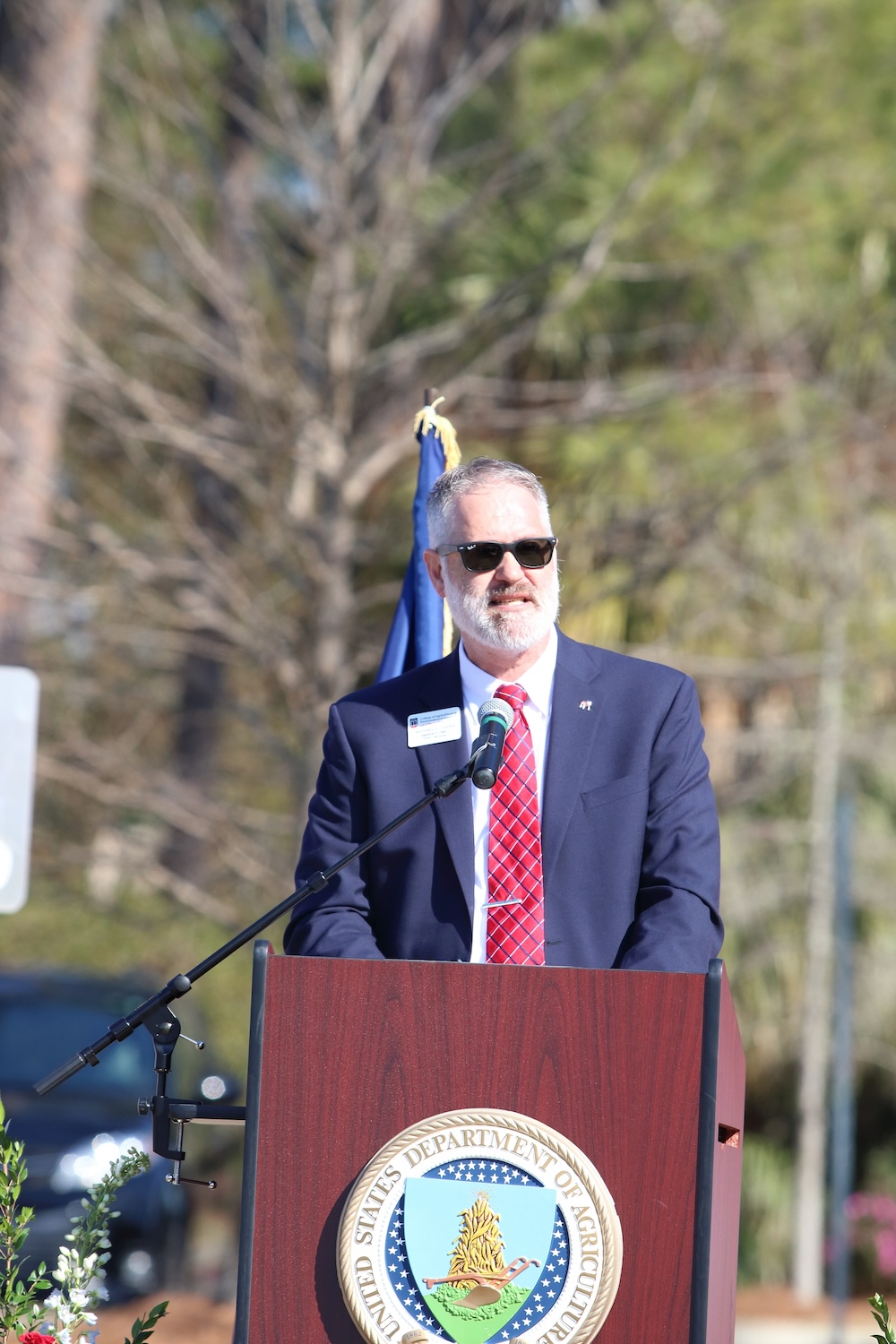 A man with a beard and sunglasses wearing a dark blue suit stands at a podium.