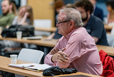Keith Kelly sits with his arms crossed in front of him at a judging table listening to student presentations during the 2024 FABricate competition. 