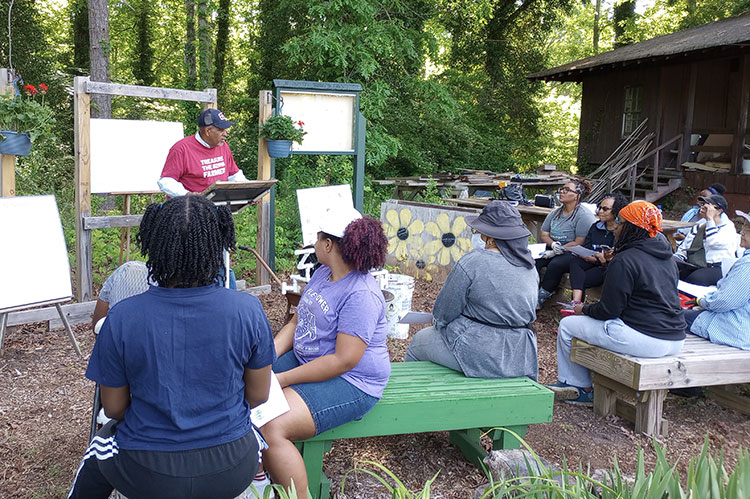 Ten community members sit on wooden benches outdoors at the GROWL Fulton County Demonstration and Teaching Garden listening to a Master Gardener Volunteer given instruction..