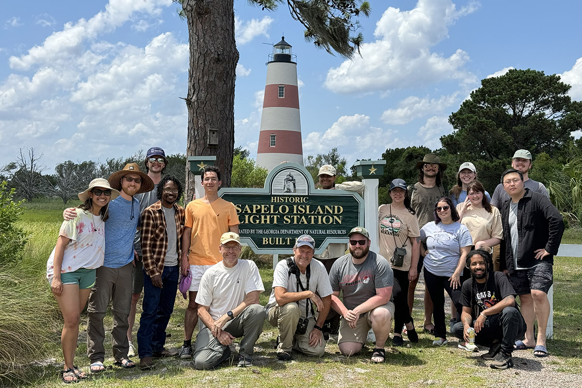 A large group of people stand around a sign that says "Sapelo Island Light Station" with a lighthouse in the backrground.