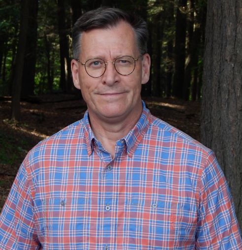 A headshot of a man wearing glasses and a blue and red plaid shirt, smiling at the camera.