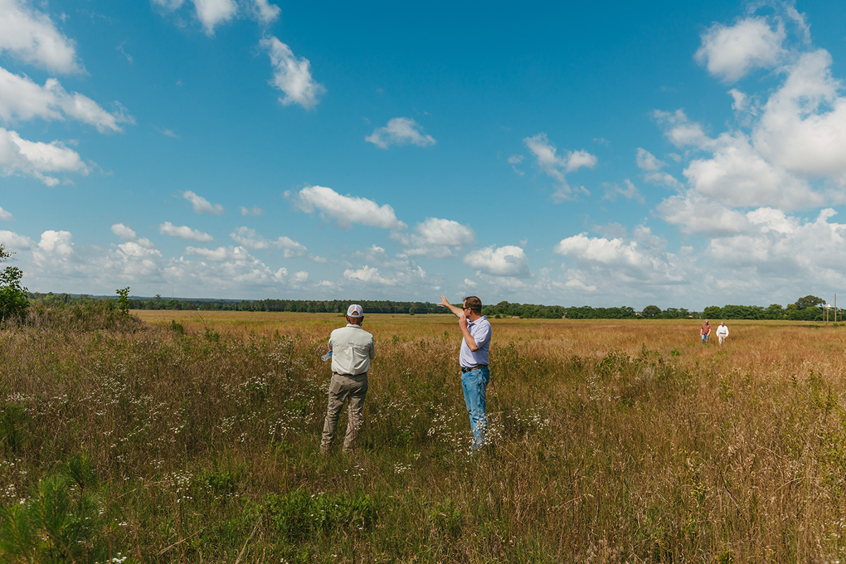 Two men stand in a large field, looking out over the land.