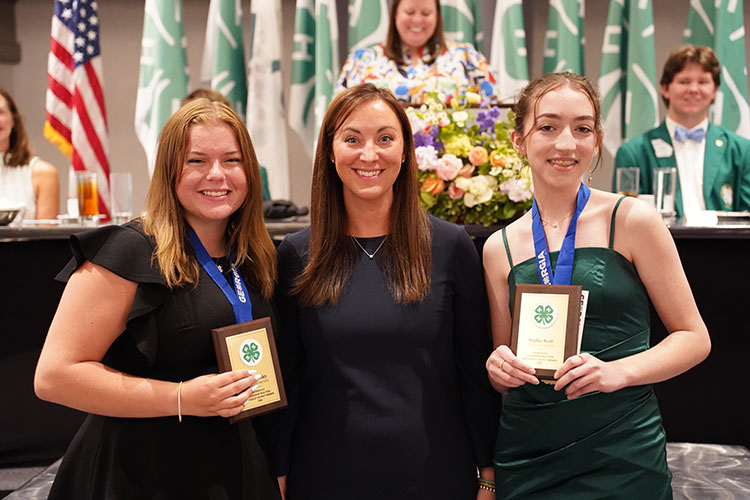 A group photo of three women, two of whom are holding plaques. The woman in the center has long brown hair and is wearing a navy blue dress. The woman on the left is wearing a black dress, and the woman on the right is wearing a dark green dress. They are standing in front of a table with floral arrangements and 4-H banners in the background.