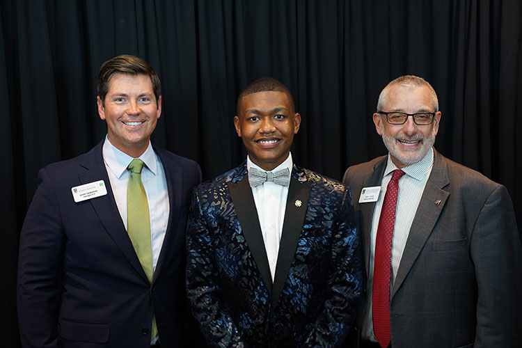 Three men posing for a photo in front of a dark curtain. The man on the left is wearing a dark suit with a light green tie. The man in the middle is wearing a dark blue patterned tuxedo jacket with a bow tie. The man on the right is wearing a gray suit with a red tie.