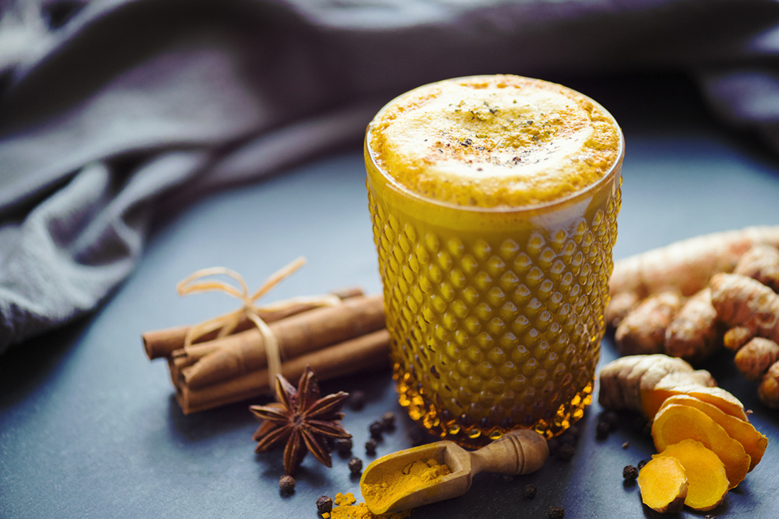 A textured glass filled with golden turmeric latte, surrounded by spices like cinnamon sticks, star anise, and turmeric roots.