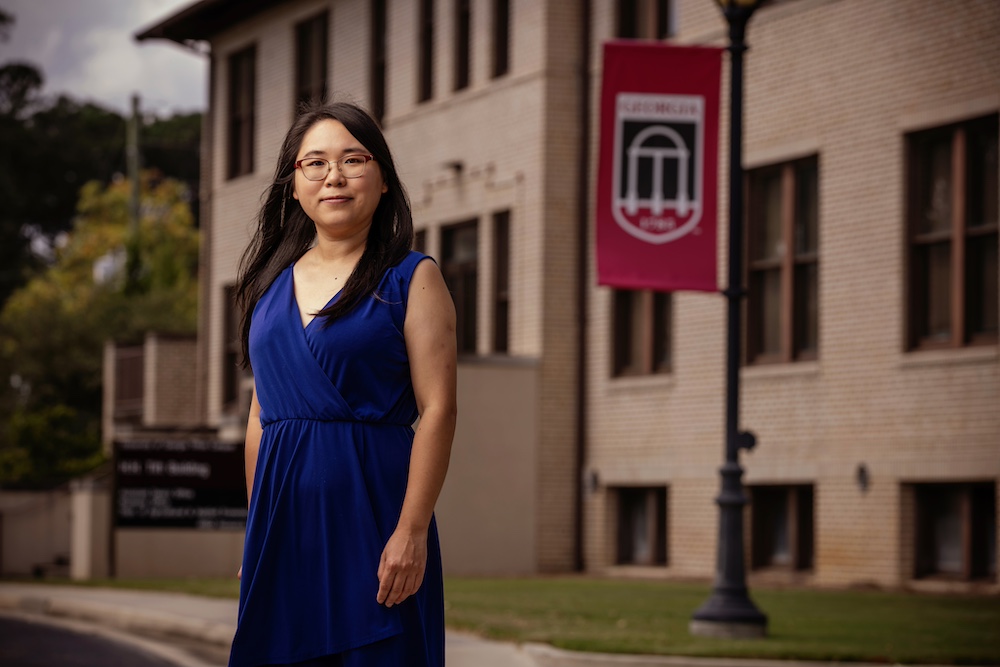 A woman with long, dark hair and glasses stands in front of a UGA building with university-branded signage