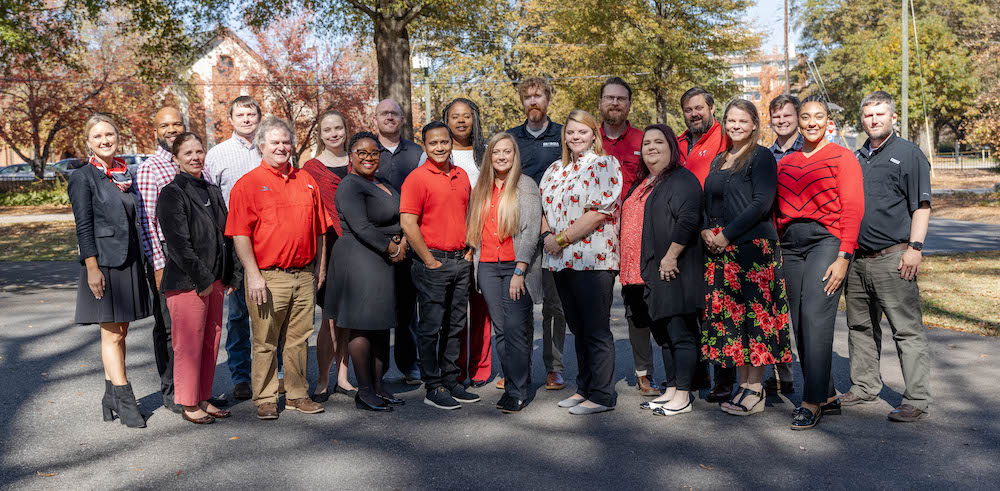 A group of 19 early and mid career professionals pose together for a photo in an outdoor setting with trees in the background. 